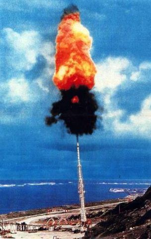 A huge gun, like an artillery cannon, fires a flaming cloud directly up into the blue sky above a coastal military base.