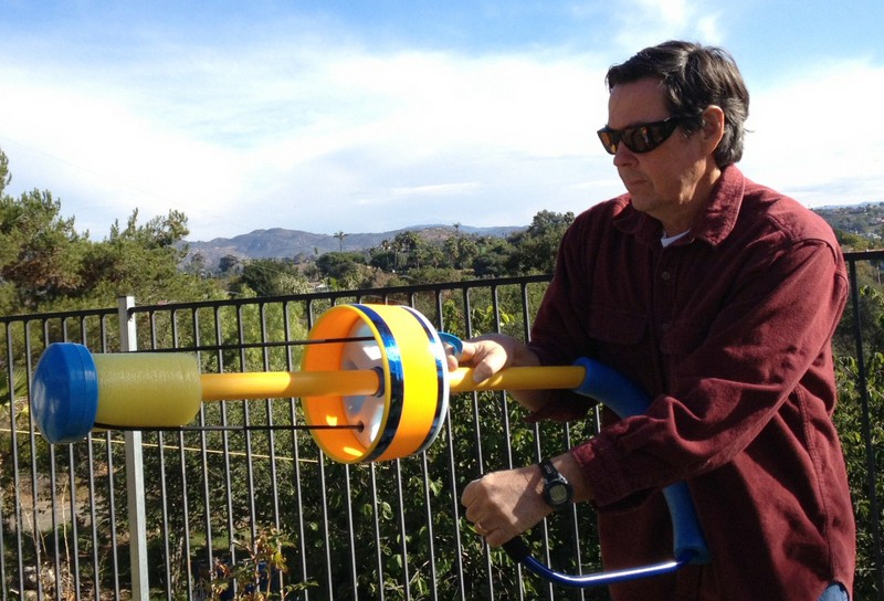 A man holds a yellow plastic rod. At the end opposite the handle is soft padding, and halfway along the shaft is a larger yellow rim.
