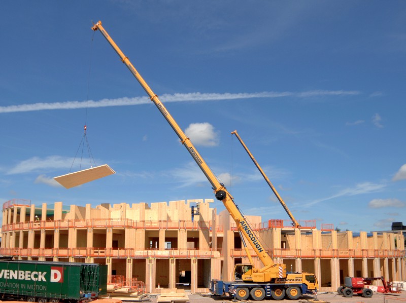 A large wooden building under construction, with a crane lifting pieces into place.