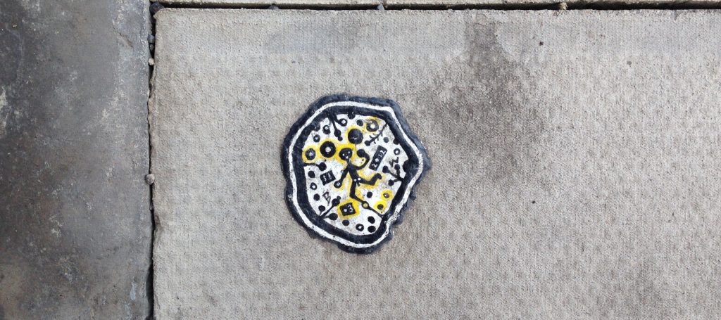 A piece of chewing gum pressed into the sidewalk. It has been colored in using marker pens to show a naked figure in the middle, smiling, surrounded by abstract squiggles.