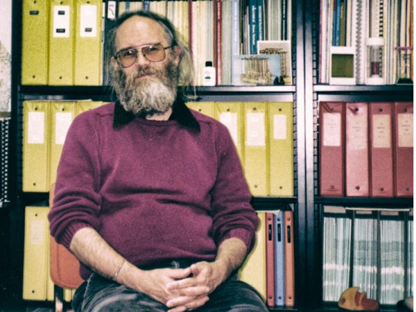 A bearded Jon Postel sits in his office in front of a large wall of binders and files.