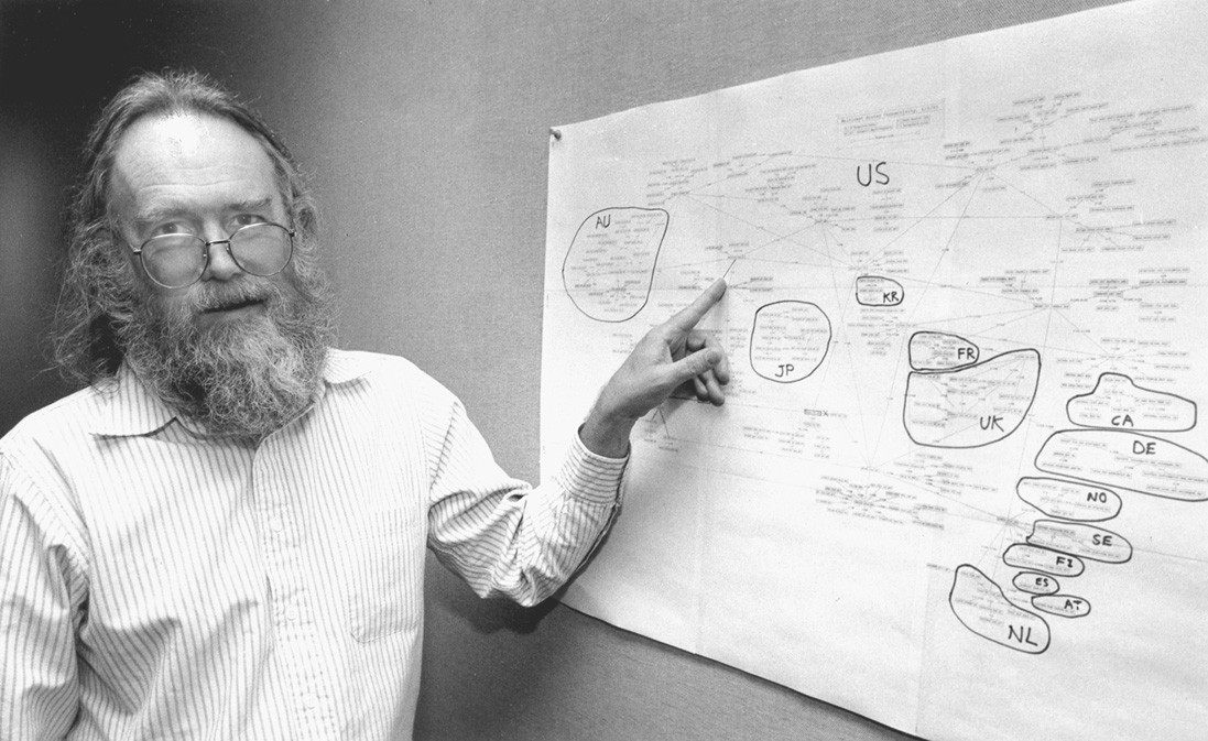 Jon Postel stands by a large piece of paper pinned to the wall, and points to it. It has a map of the internet on it as of 1994.