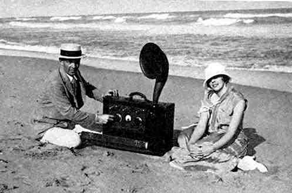 A man and a woman sit besides a large portable radio unit on the beach.