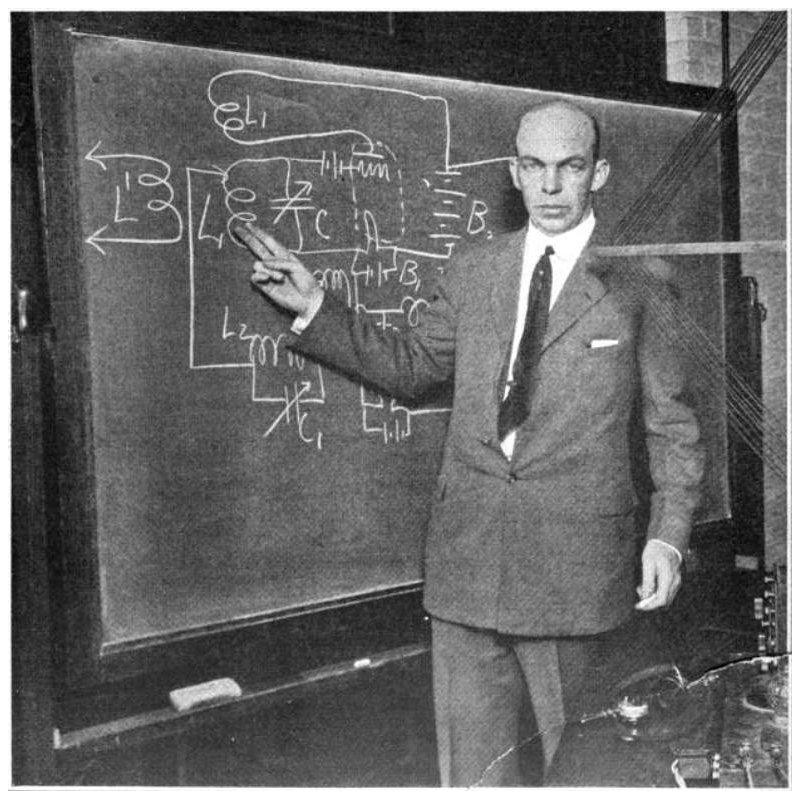 A man stands at a blackboard, pointing at the circuit diagram drawn on it.