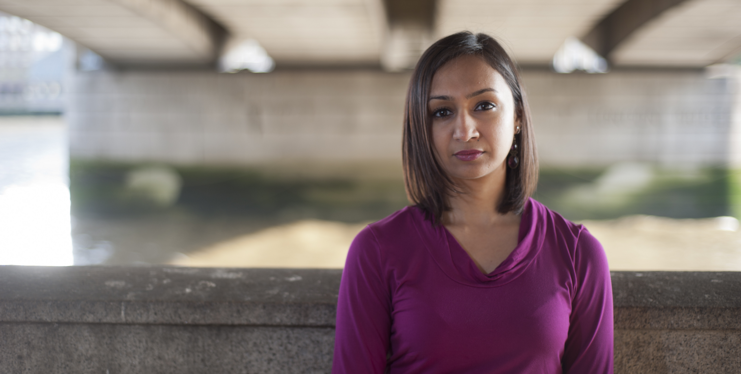 A woman in a purple shirt poses for the camera beneath a bridge.