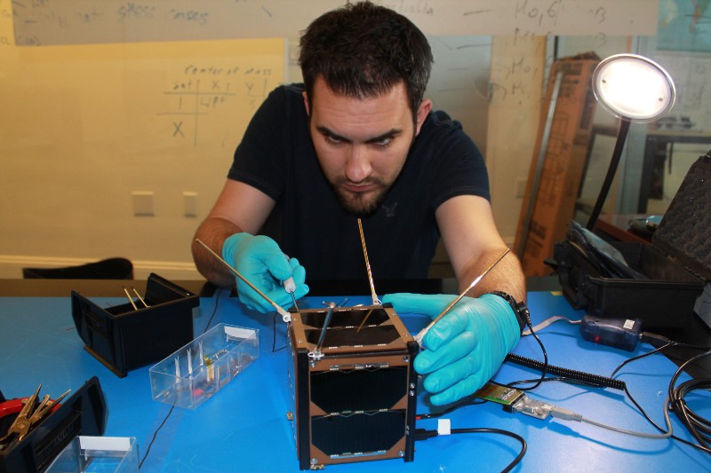 A man wearing blue latex gloves delicately works on one of the cube-shaped satellites, which is only about the same size as a toaster.