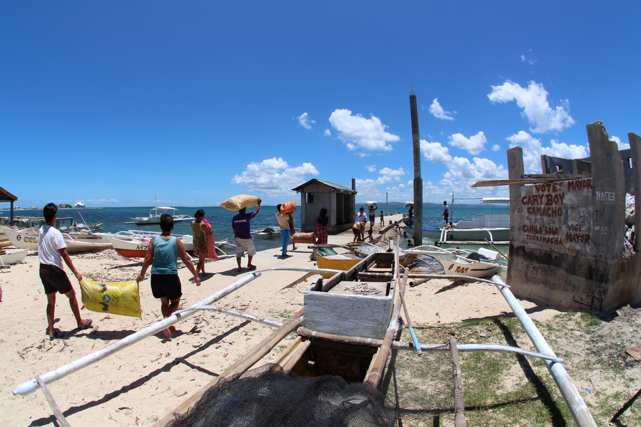 A group of people load sacks onto small boats at a beachside dock.