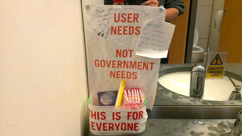A rudimentary paper bucket containing sanitary pads and tampons. The sign reads: "User needs, not government needs."