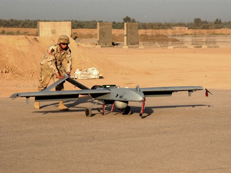 A soldier pushes a drone along the ground in a sandy desert-looking area.