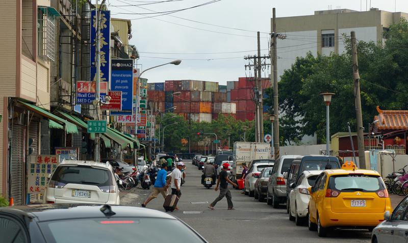 A street near the port. Cars and other vehicles line both sides, there are stores and pedestrians, and at the end of the street is a wall of container ships where the port is.