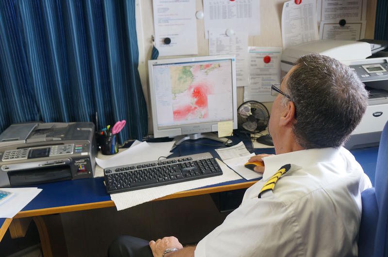 The captain sits at his desk in his office, looking at weather forecasts on his computer.