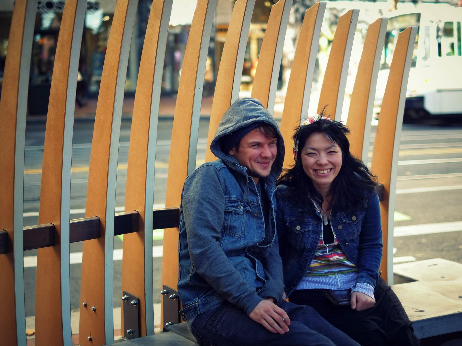 Two people sit on a bench in front of a wooden fence.