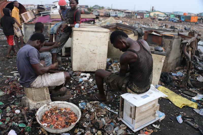 A group of men sit and stand around in a garbage dump site, going through the waste and collecting items worth reselling.