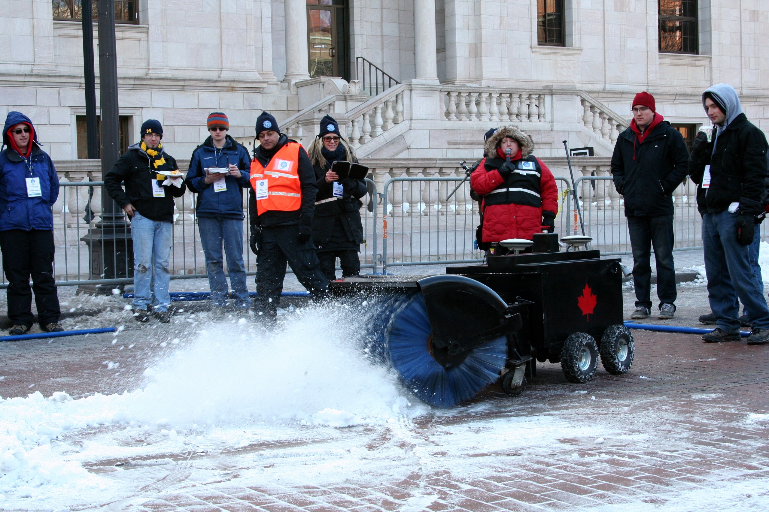 An autonomous snow sweeping machine moves through a town square, while a crowd watches.