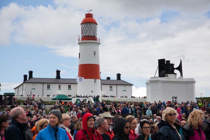 A large crowd gathered at an event, near a red and white lighthouse.