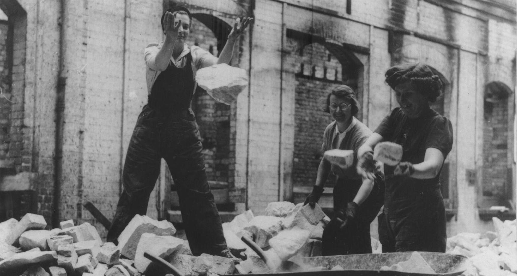 Three women wearing work clothes and gloves carry large chunks of stone around a building site.