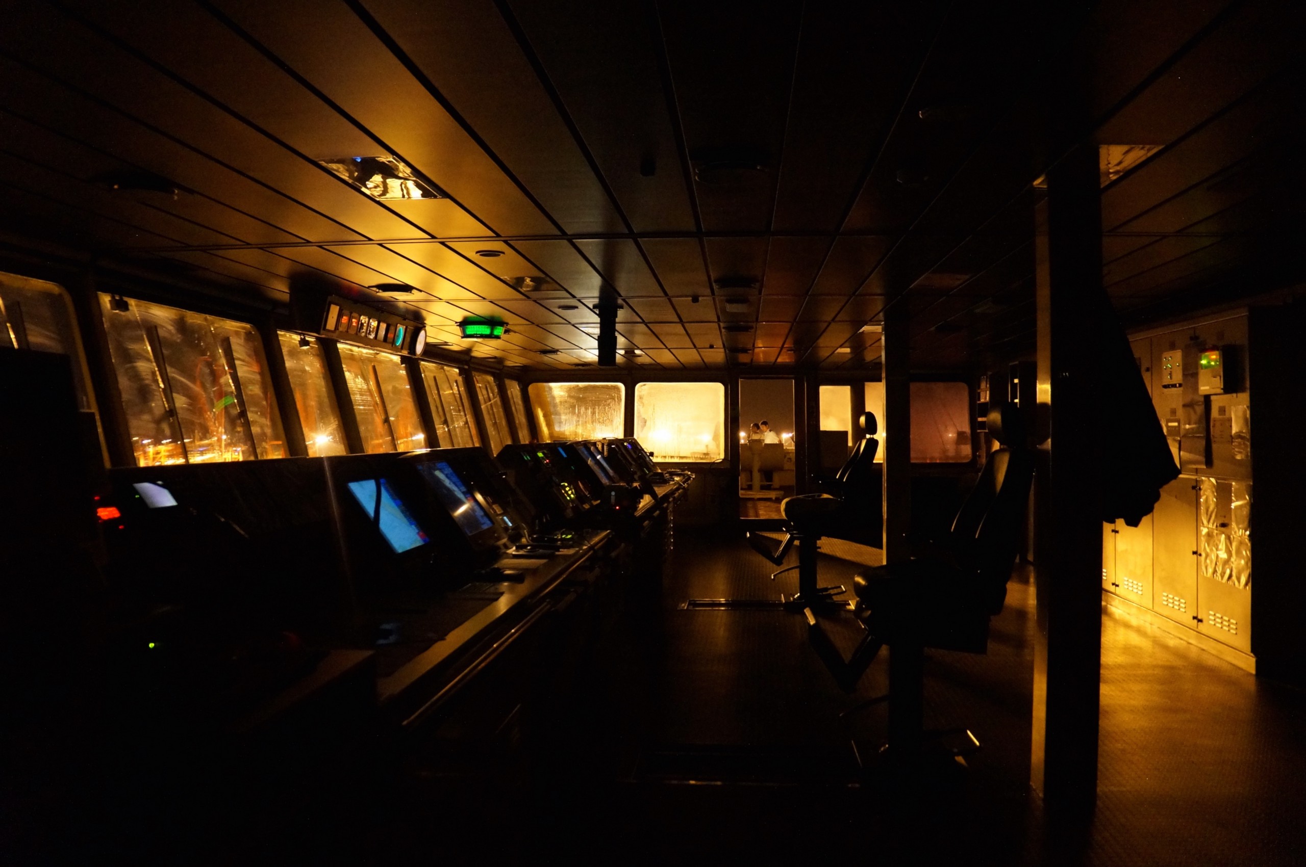 The bridge of a container ship. Many computer screens line a console against the forward-facing window, along with radio and other communications equipment.