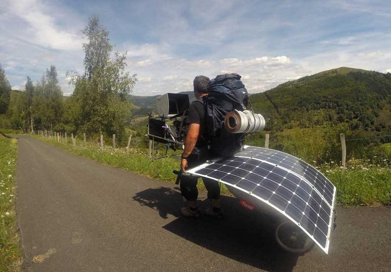 A man carrying a backpack, pulling a solar panel on wheels behind him, with a computer monitor mounted in front of him.