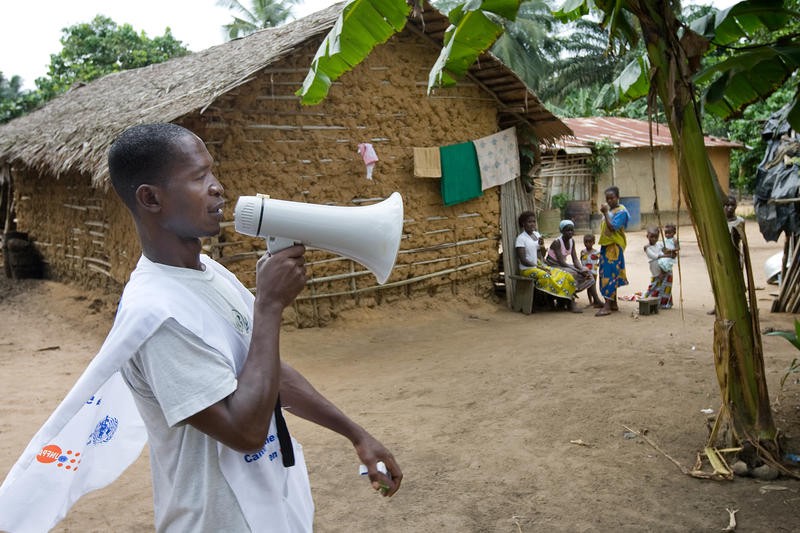 A man with a bullhorn walks around a village, speaking into it.
