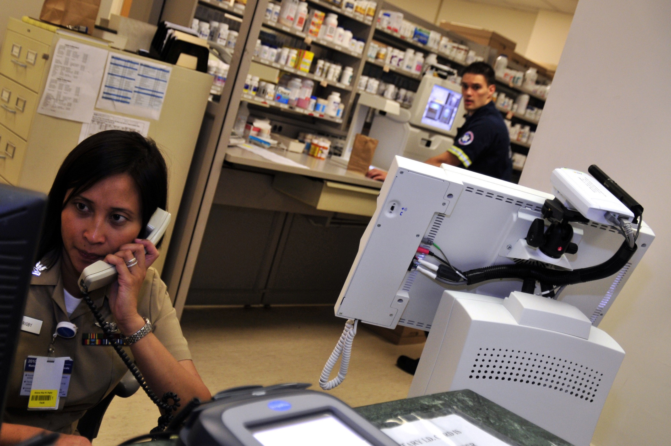 A woman speaks on the phone in a pharmacy while a man in EMT uniform looks on.