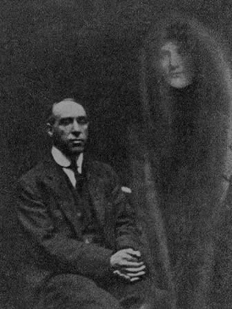 Harry Price sits, left, with a misty figure standing in a robe and hood to the right.