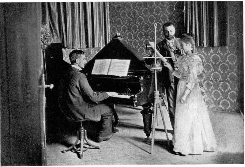 A pianist and a singer perform for a microphone, held by another man. They are dressed formally.