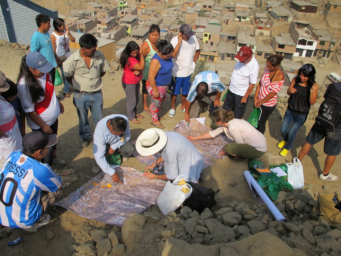 A group of people standing on a hillside surround some maps on the ground, which are being pointed at and examined. In the background, the favela can be seen rolling down the hillside.