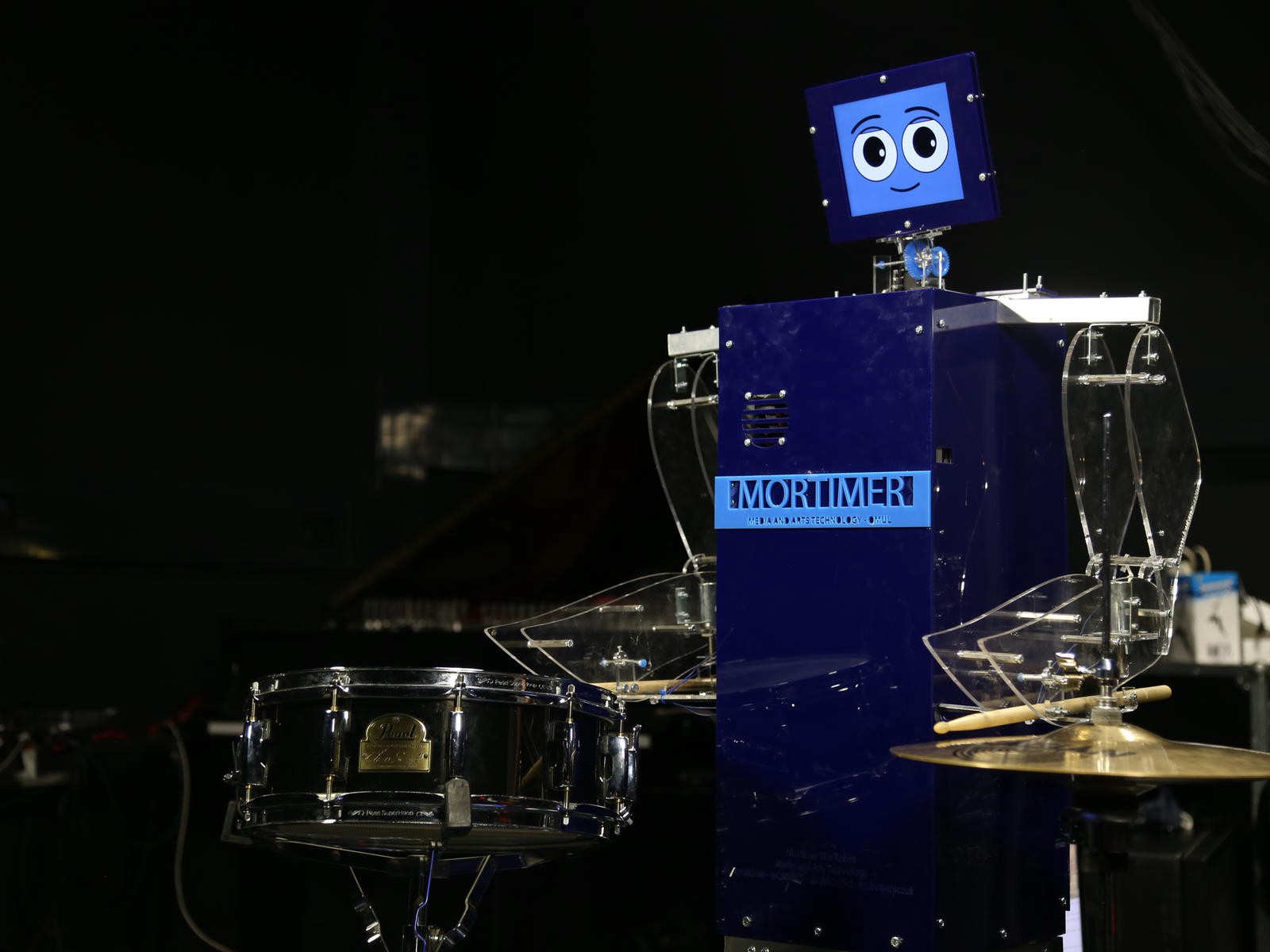 A large blue box with arms that are holding drumsticks. On top of the box is a smaller box, acting as a head. There is a smiling face on the head.