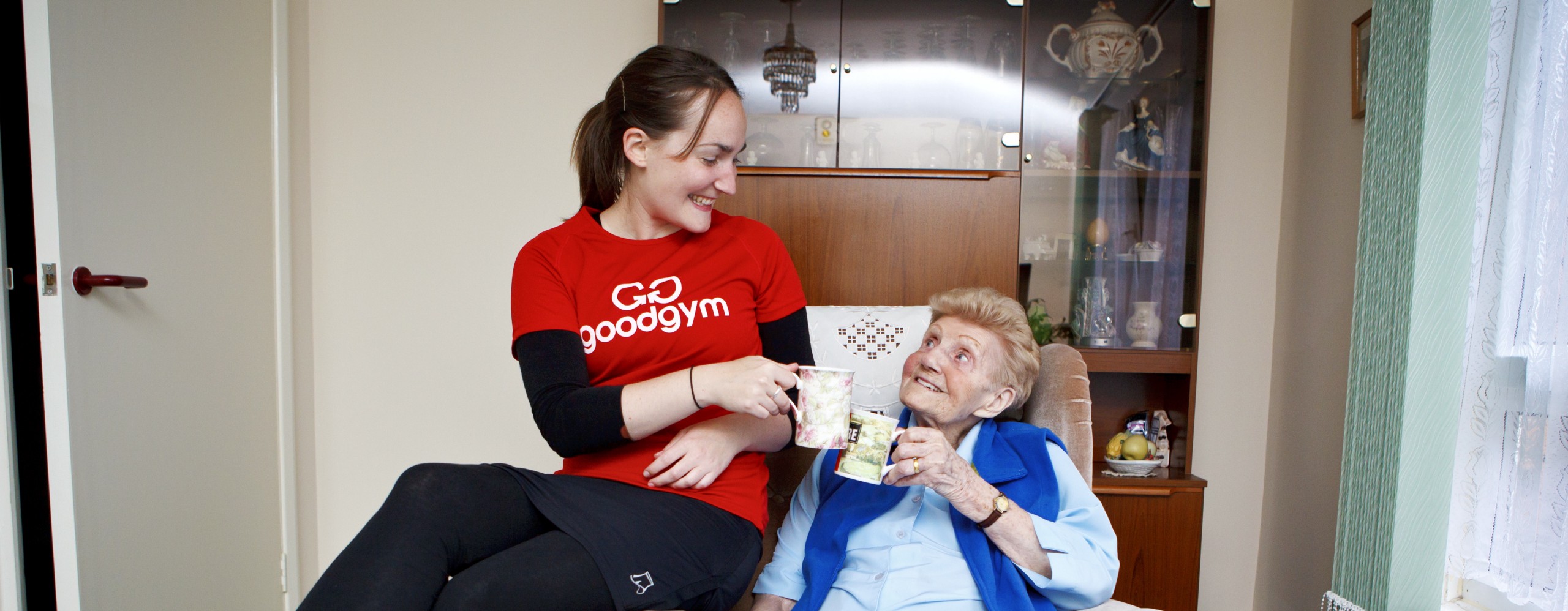 Two women–one older in a chair, one younger and wearing a GoodGym t-shirt–drink tea together.