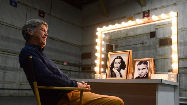 Steven Johnson sits in a makeup chair in front of a lighted mirror. On the table is a framed picture of Hedy Lamarr.