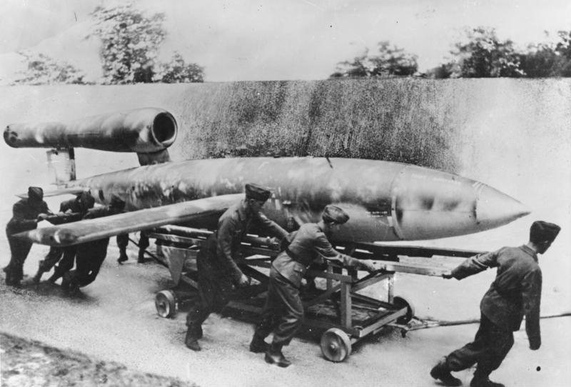 Soldiers wheel a V1 rocket on a trolley down a runway.
