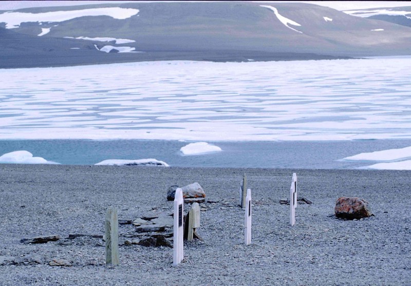 Three basic graves in a line on a gravel beach, with ice out over the water and bare hills in the distance.