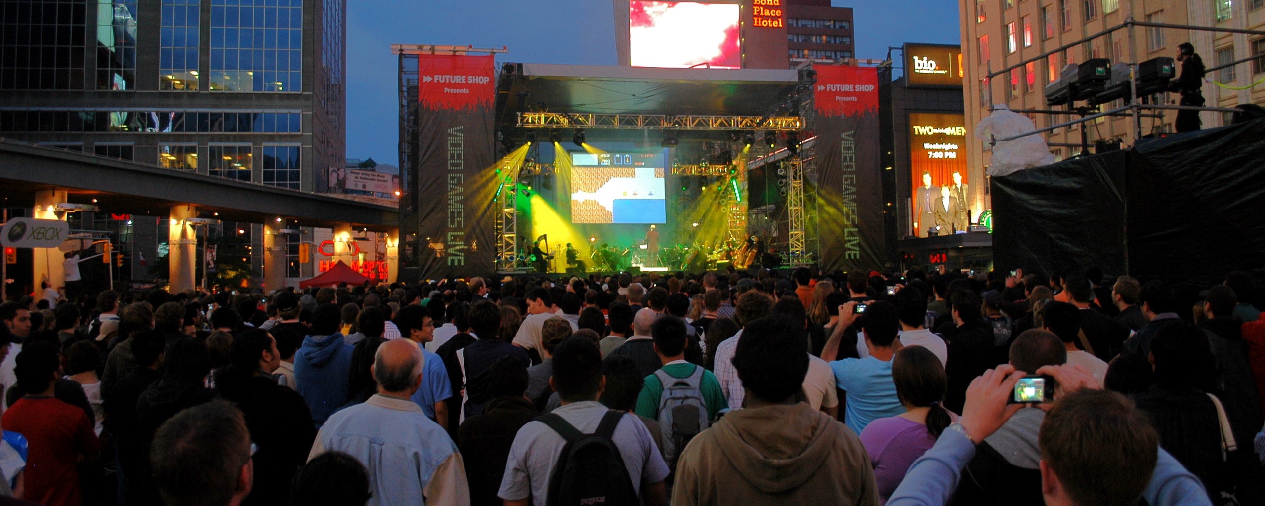 A crowd of people in an outdoor city square watch an orchestra perform on a stage, in front of a projection of a level from The Legend of Zelda.