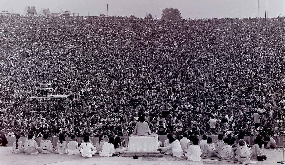 A view of the huge crowd at Woodstock from behind the stage, looking out over the heads of the performers. The crowd takes up the entire field of view, to the horizon.