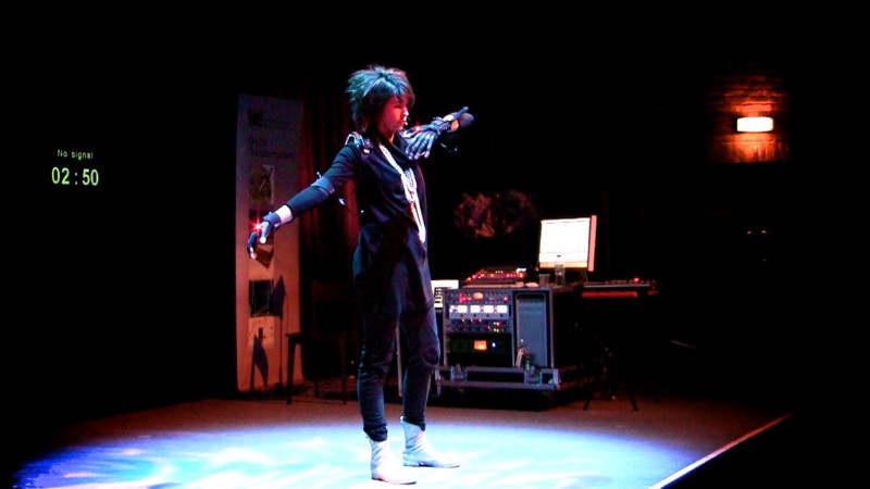 The artist Imogen Heap stands on stage in a specific pose, as she uses her physical positions to perform music.