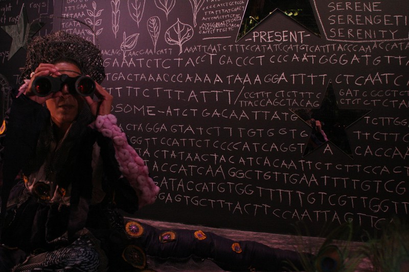 A woman sits in front of a blackboard covered in written letters. She looks through binoculars.