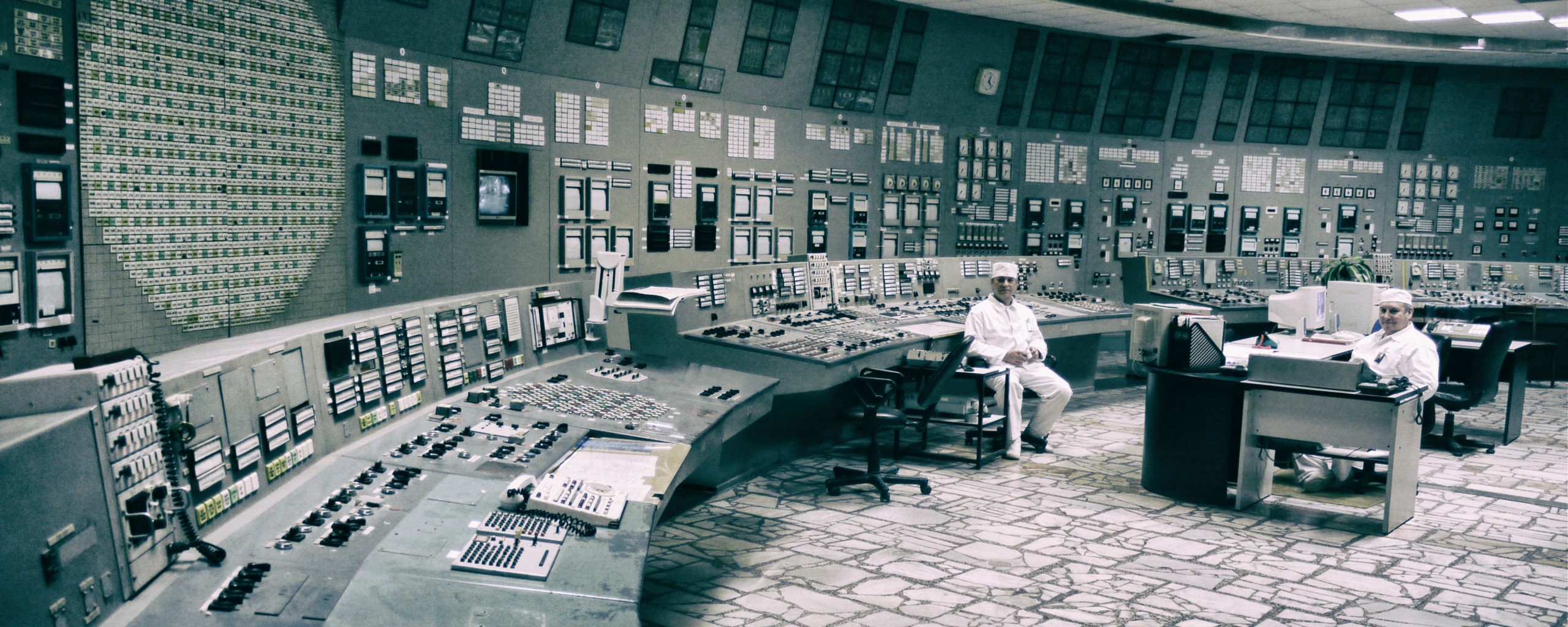 Two men in white suits and caps sit in a large room with a huge control panel displaying innumerable switches, buttons, displays, and lights.