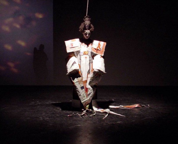 A woman stands on a stage in darkness, wearing an elaborate costume.