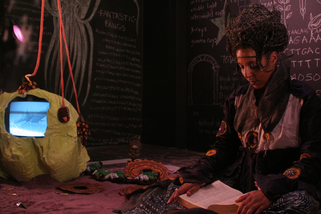 A woman sits reading in a room, next to a TV. The walls are blackboards covered in writing.