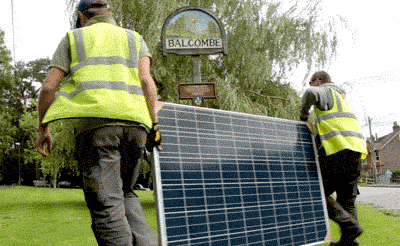 Two men carry a solar panel past the sign at the entrance to the village of Balcombe.