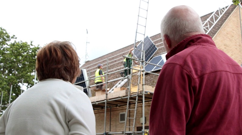Two people in the foreground watch two men on the roof of a building, attaching solar panels.
