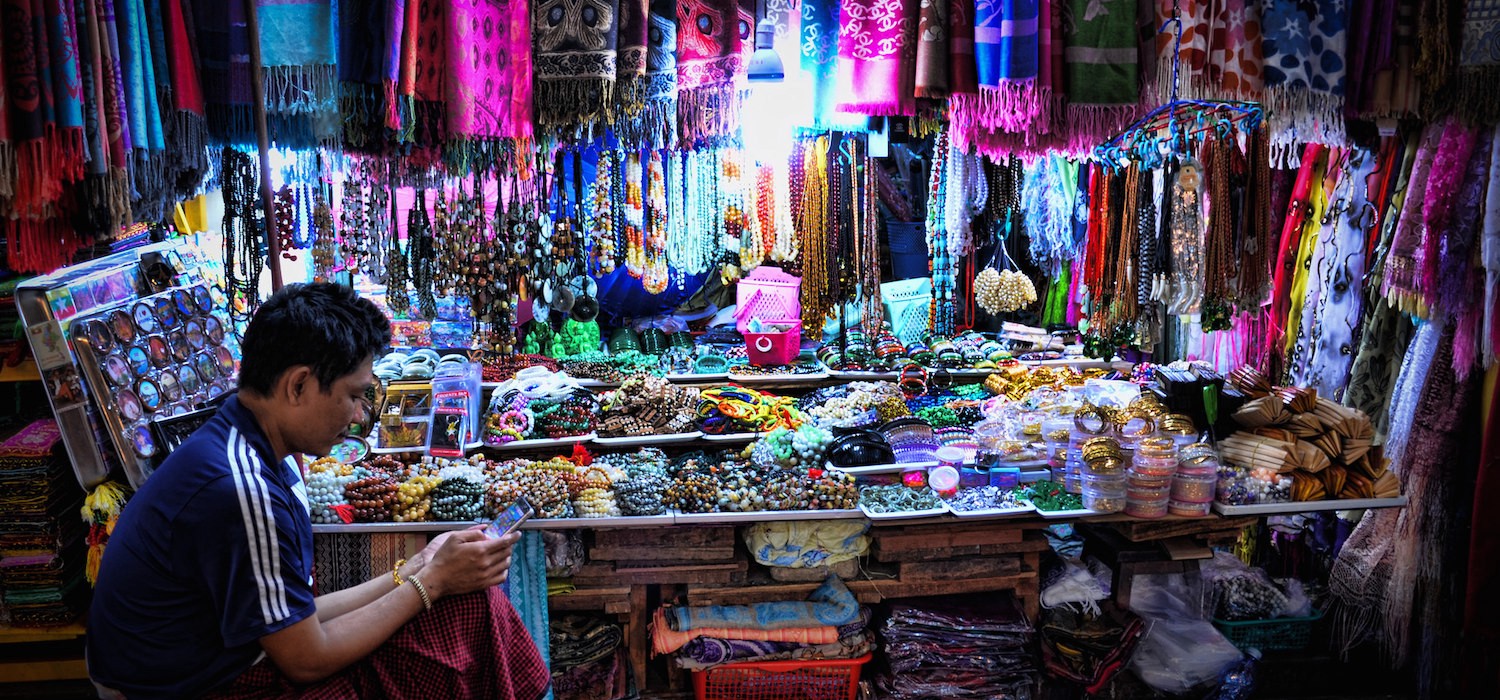 A man sits at a market stall with jewelery for sale. He plays on his phone.
