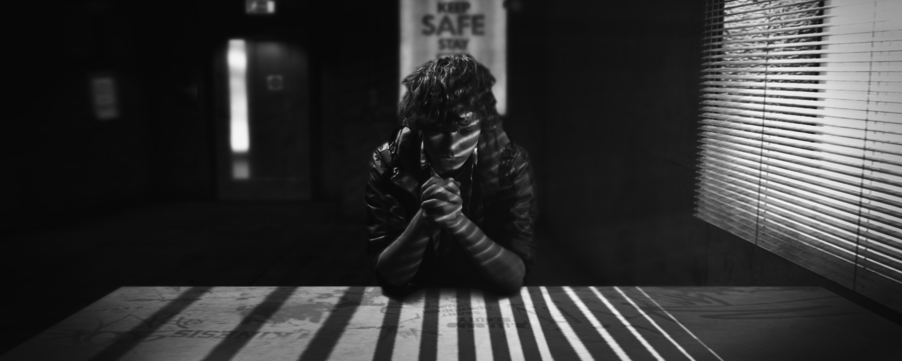 A person sitting with their hands clasped at a table in a dark room with the shutters down. The light is casting shadows on them.