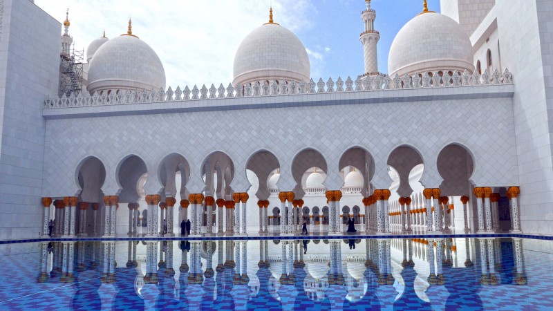 The exterior of a white marble mosque with a pool of water outside.
