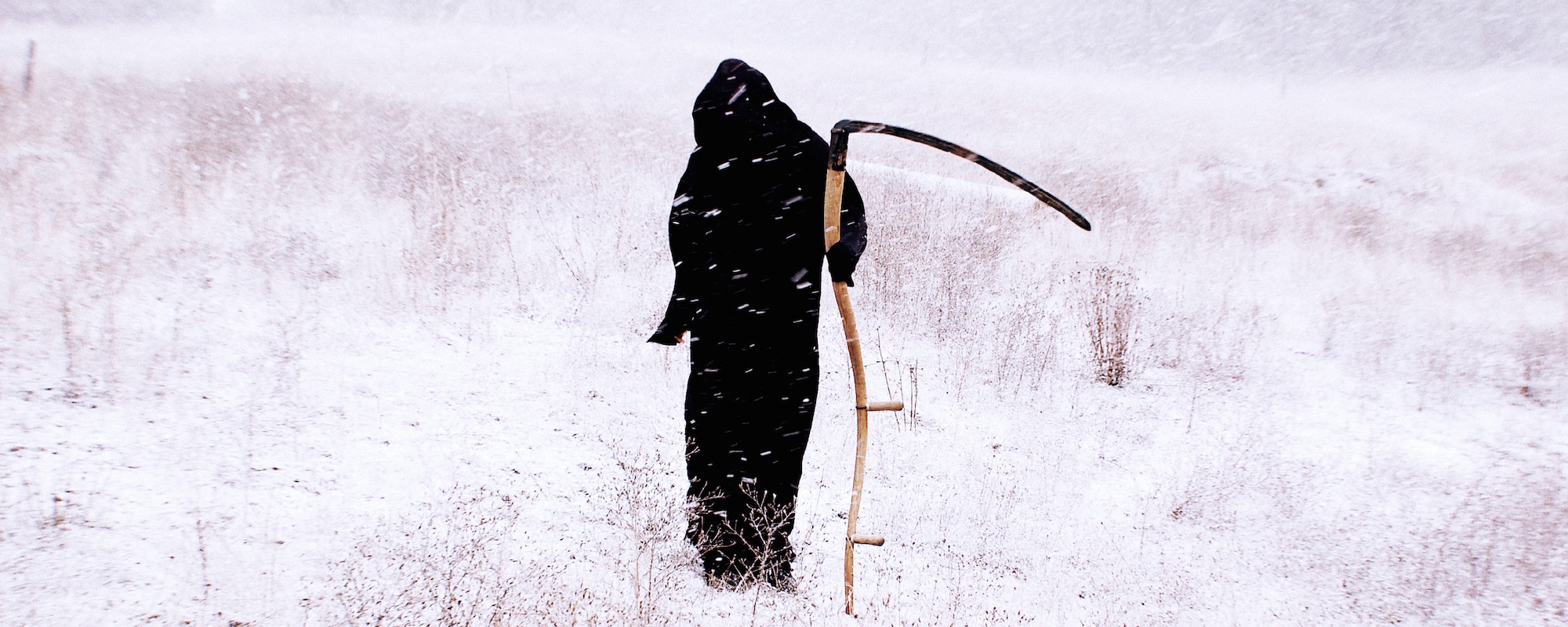 A person dressed as death, in a long black hooded robe with a scythe, stands in a snowy field.
