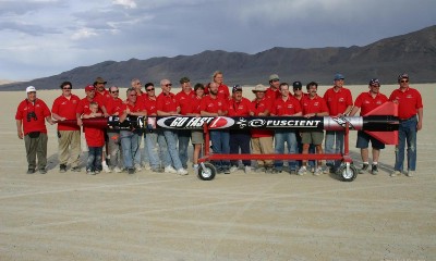 A large group of people in red shirts collectively holds, horizontally, a black and red rocket, while standing on a flat desert basin.
