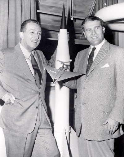 Walt Disney stands next to Werner von Braun. Disney is leaning on a model rocket the same height as himself, while von Braun holds a small model passenger spaceship in his right hand.