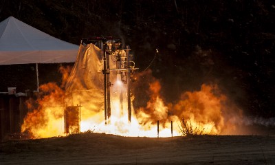 A naked rocket engine attached to a stand just above the ground in an outside area fires, blasting fire in all directions.