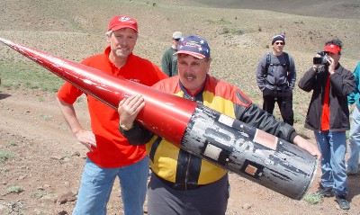 Ky Michaelson, wearing a button-down shirt and a baseball cap, holds the top of the GoFast rocket–it is approximately five-feet long, and a foot wide.