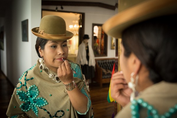 An Aymara woman in a bowler hat and traditional dress puts on lip liner.